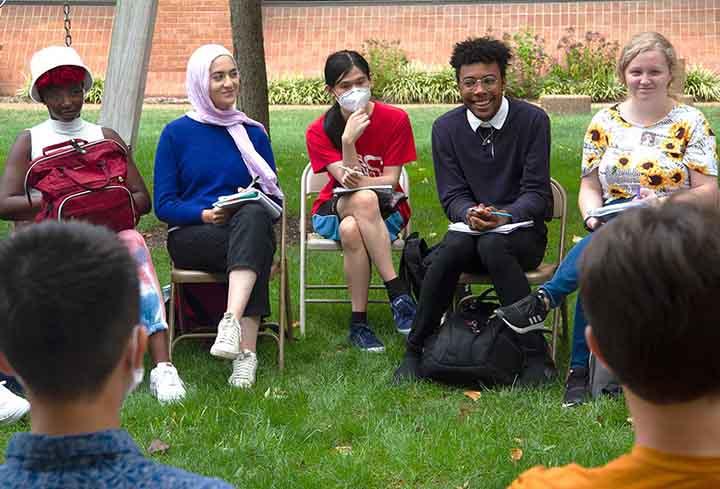 A group of students sit on folding chairs set up in a circle on a lawn for discussion